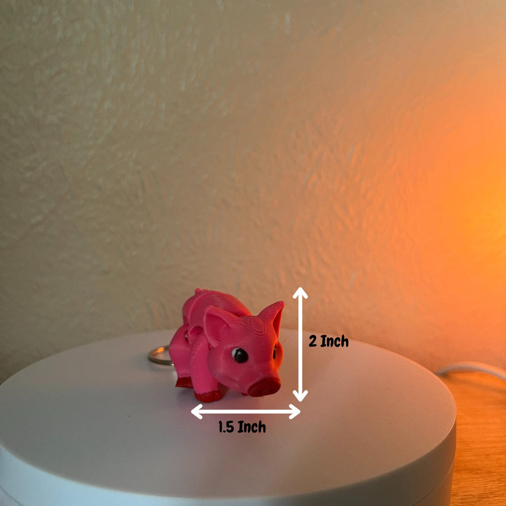 A pink pig keychain, measuring 2 inches tall and 1.5 inches wide. The pig has black eyes and a red snout and hooves. The keychain is on a white surface.