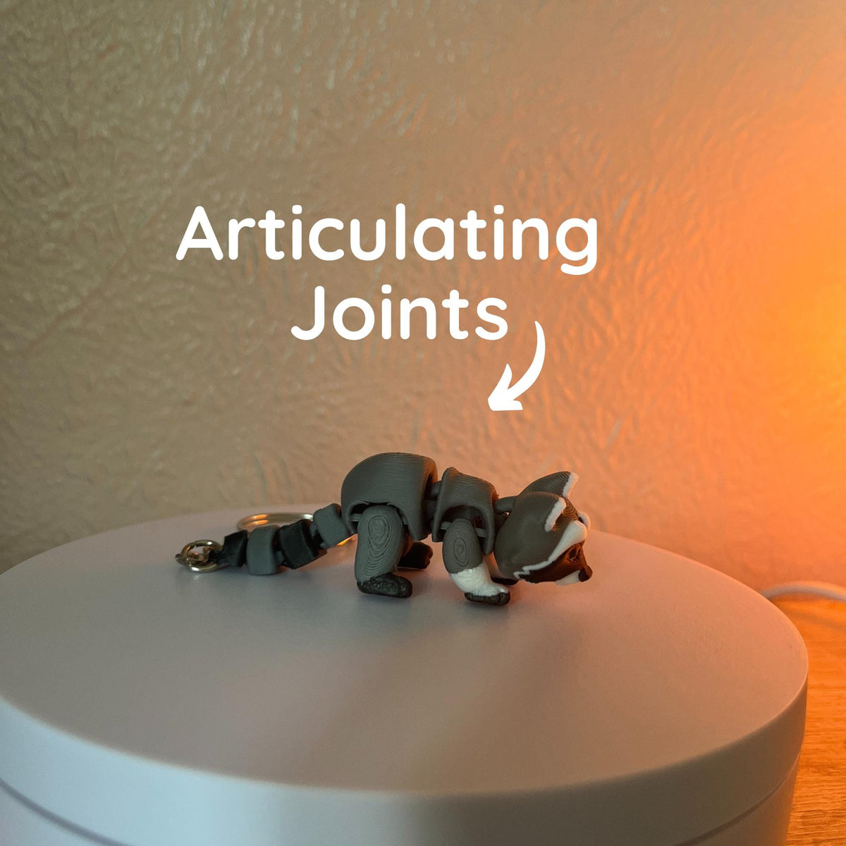 A gray and white articulated raccoon figurine with a keychain attachment. The text "Articulating Joints" is above the raccoon. The raccoon is on a white surface.