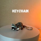 A gray and white raccoon keychain with a gold-colored keyring. The word "KEYCHAIN" is displayed in white block letters above the raccoon figurine. The raccoon has a black mask, gray body, and white paws.