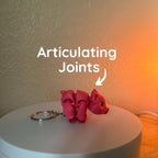 A pink articulated pig keychain. The pig has a segmented body with visible joints, allowing for movement. The keychain includes a silver-colored ring. The text "Articulating Joints" is displayed above the pig.