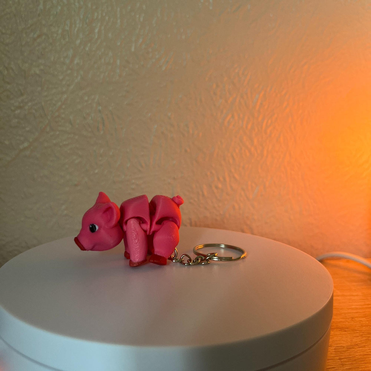 A pink pig keychain with a silver keyring. The pig is made of a folded material, with a small tail and a detailed face. The keychain is on a white surface.