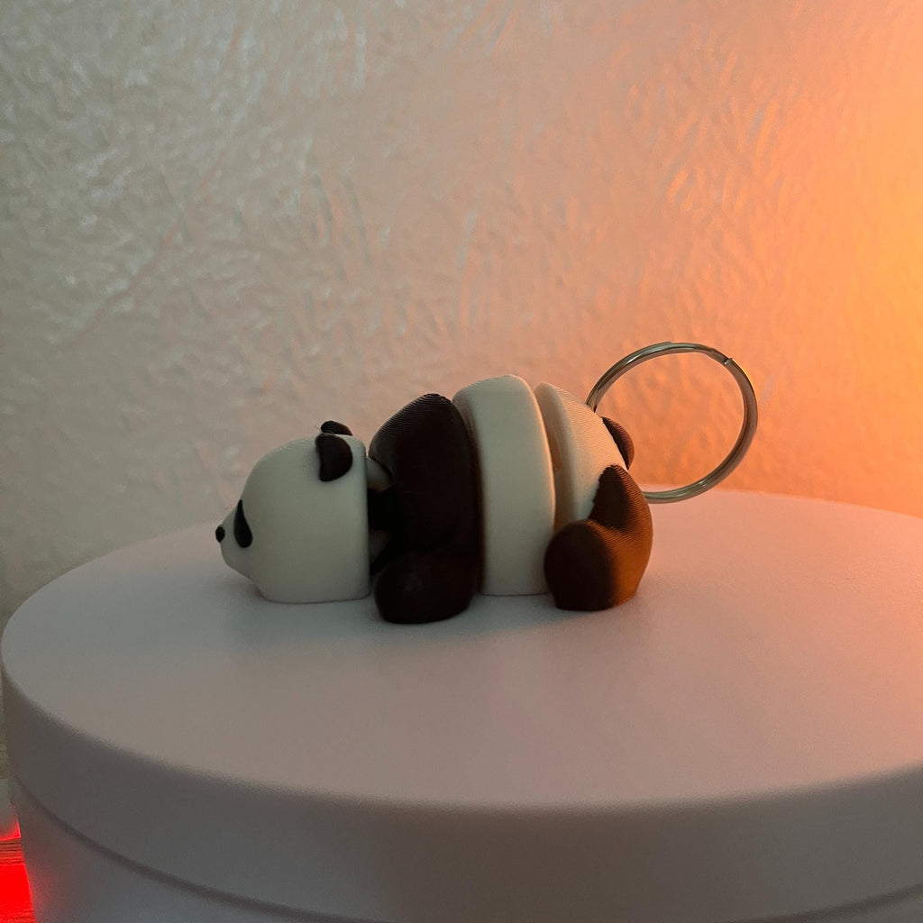 A panda bear keychain in a lying down position. The panda has a white body with black accents on the ears, eyes, and limbs. A silver-colored metal ring is attached to the panda's back.