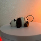 A panda bear keychain in a lying down position. The panda has a white body with black accents on the ears, eyes, and limbs. A silver-colored metal ring is attached to the panda's back.