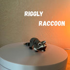 A small, gray and black raccoon figurine with white accents, lying on a white surface. The text "RIGGLY RACCOON" is displayed above the figurine. The figurine has a keychain attachment.