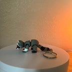 A gray raccoon figurine with a white face and paws, and a brown tail. The raccoon is attached to a gold-colored keychain. The figurine is made of a hard material and is lying on a white surface.
