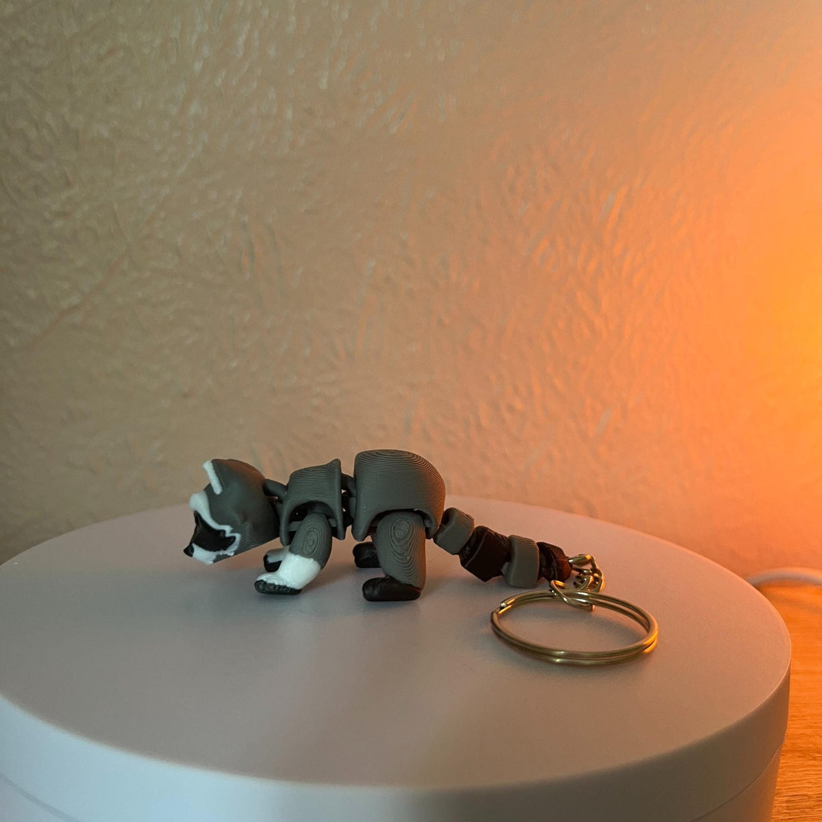 A gray raccoon figurine with a white face and paws, and a brown tail. The raccoon is attached to a gold-colored keychain. The figurine is made of a hard material and is lying on a white surface.
