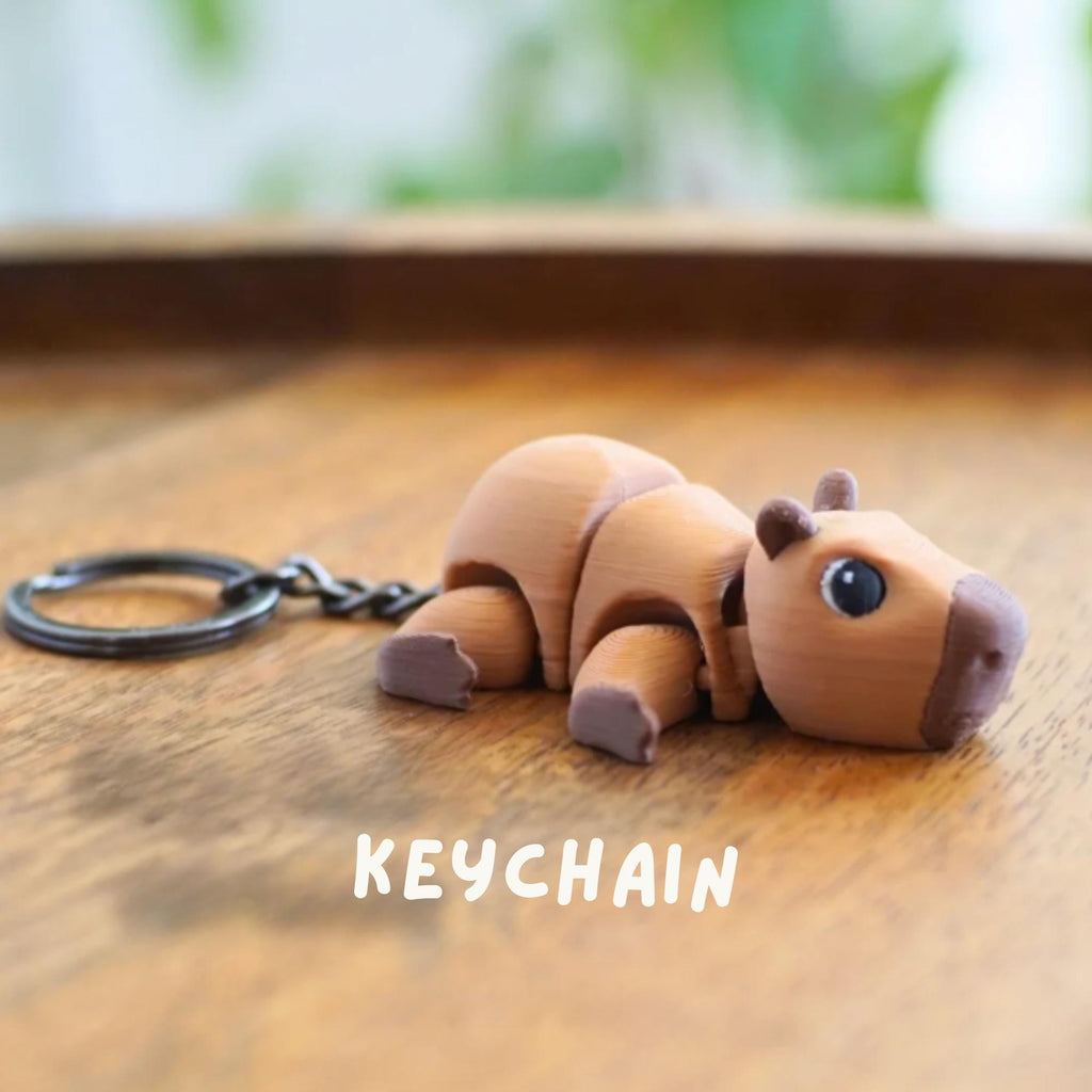 A brown capybara keychain with a dark metal keyring and chain. The keychain is lying on a wooden surface. The word "KEYCHAIN" is printed in white below the capybara.