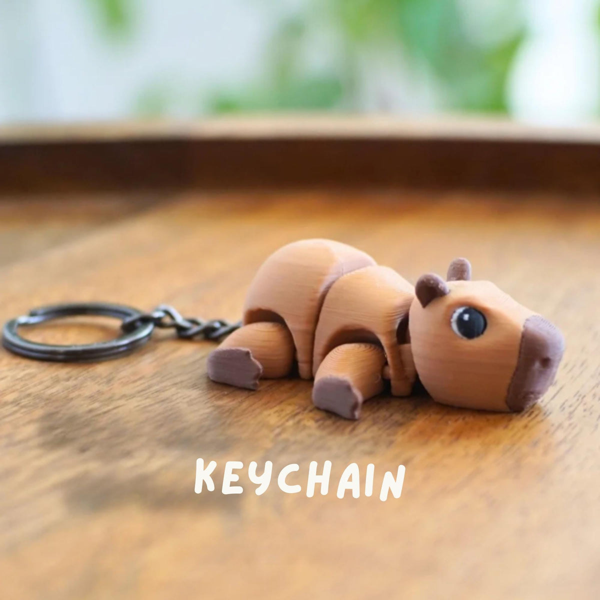 A brown capybara keychain with a dark metal keyring and chain. The keychain is lying on a wooden surface. The word "KEYCHAIN" is printed in white below the capybara.