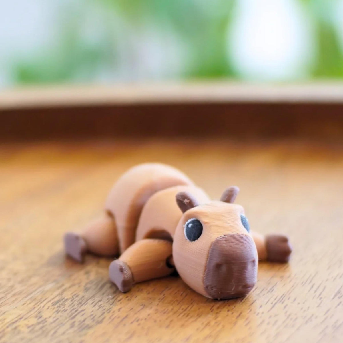 A 3D-printed capybara figurine in shades of brown, lying on a wooden surface. The toy has a detailed face with dark eyes and a dark brown nose. The capybara's body is segmented, and its legs are extended.