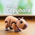 A tan and brown articulated capybara toy with the text "Clumsy Capybara" in white. The toy has a detailed design with visible joints and a dark brown nose. The background is blurred, suggesting a natural setting.