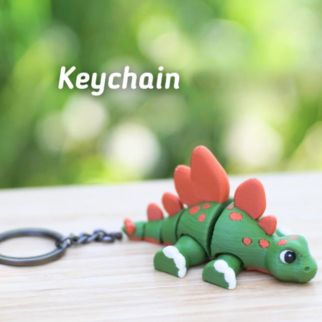 A green and orange stegosaurus keychain with the word "Keychain" in white text. The dinosaur has red plates on its back and white accents on its feet. The keychain is attached to a metal ring.