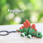 A green and orange stegosaurus keychain with the word "Keychain" in white text. The dinosaur has red plates on its back and white accents on its feet. The keychain is attached to a metal ring.