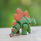 A green and orange stegosaurus toy with articulated joints. The dinosaur has orange plates along its back and tail, with orange spots on its body. The toy's feet are white, and it is posed on a wooden surface.