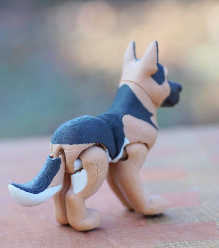 A 3D printed German Shepherd dog figurine featuring a black and tan coat with white accents on the tail and legs. The figure displays visible horizontal layer lines from the printing process and has articulated joints at the tail and legs.